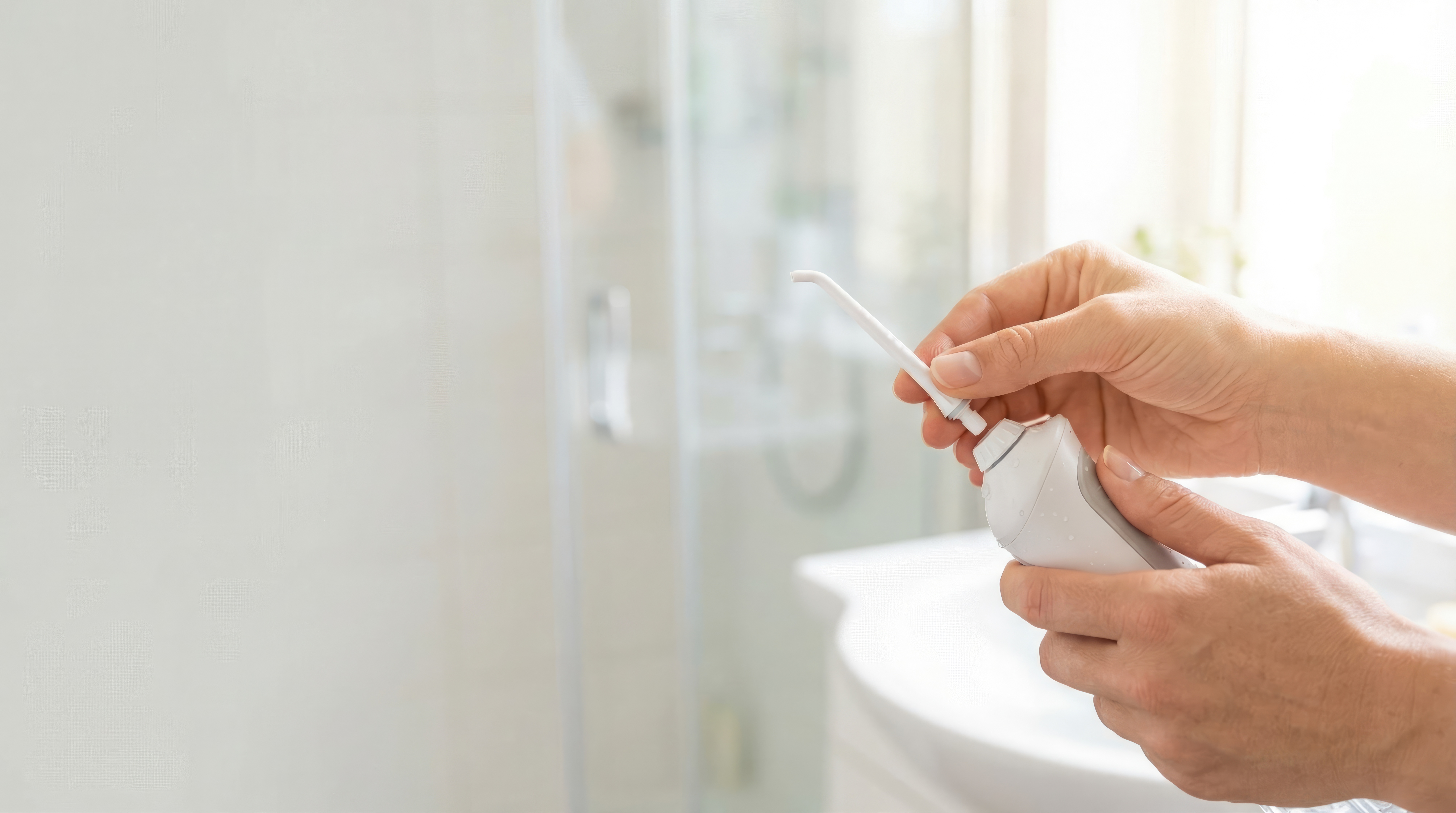 A person's hands are shown carefully inserting the nozzle into a white water flosser in a bright, modern bathroom setting.