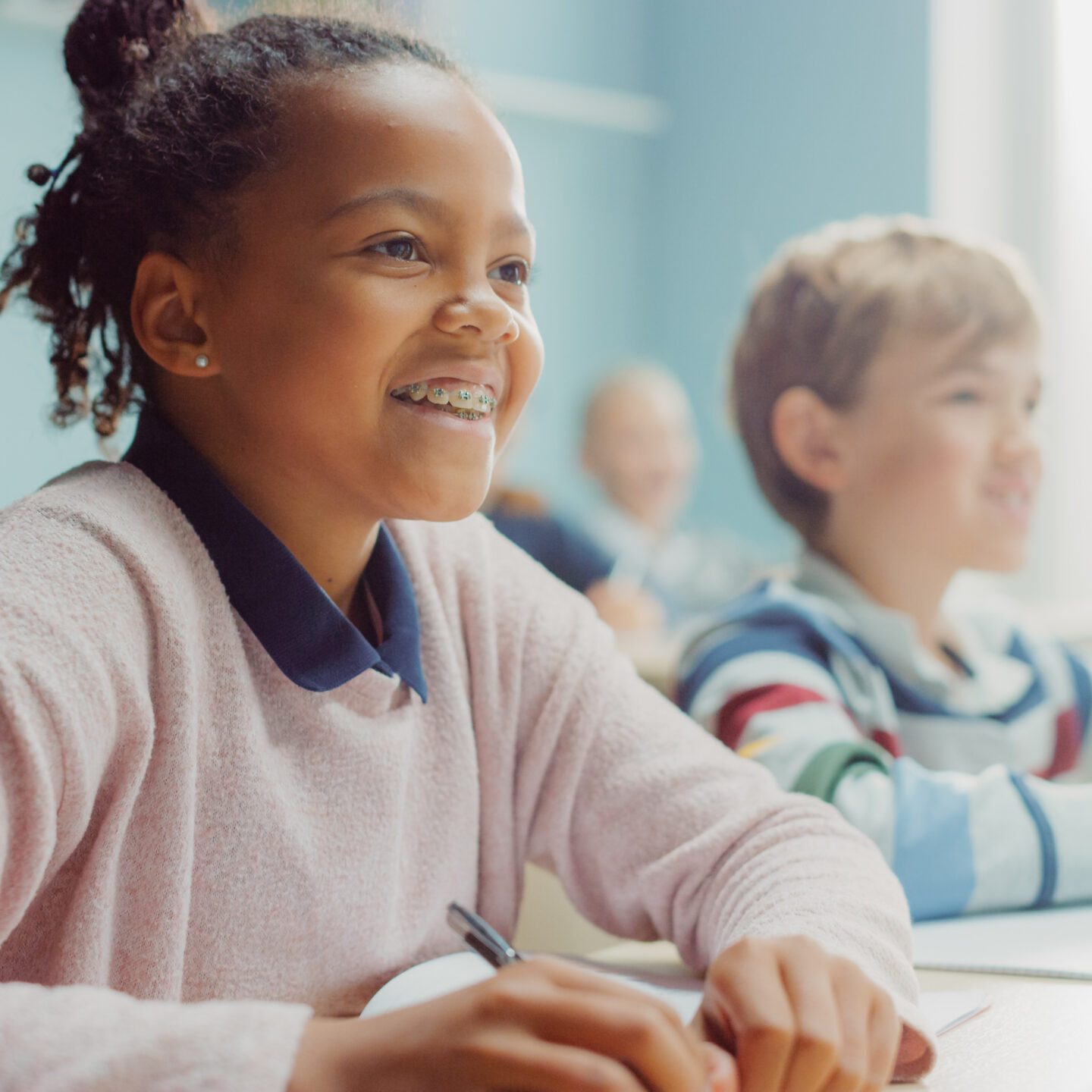 In Elementary School Class: Portrait of a Brilliant Black Girl with Braces Writes in Exercise Notebook, Smiles. Junior Classroom with Diverse Group of Children Learning New Stuff